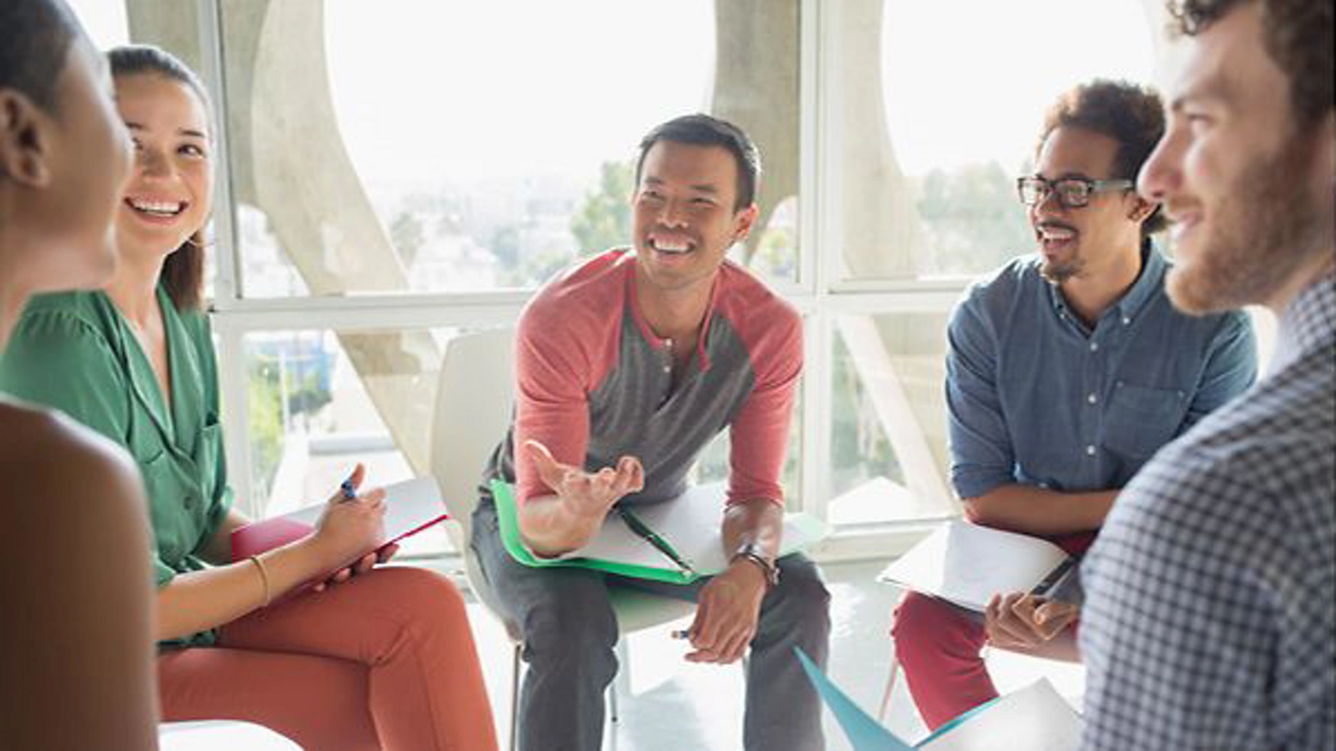 A group of diverse individuals engaged in a collaborative discussion in a bright, modern office setting. The participants are seated in a semi-circle, holding notebooks and pens, suggesting an interactive brainstorming session. Large windows in the background allow natural light to illuminate the space, creating a professional yet relaxed atmosphere.