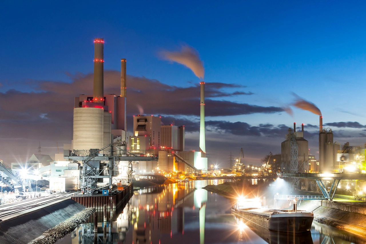A coal power plant sits at the river's edge at dusk.  Lights from the buildings and streets glow, and a reflection of the whole scene can be seen on the water of the calm river below.