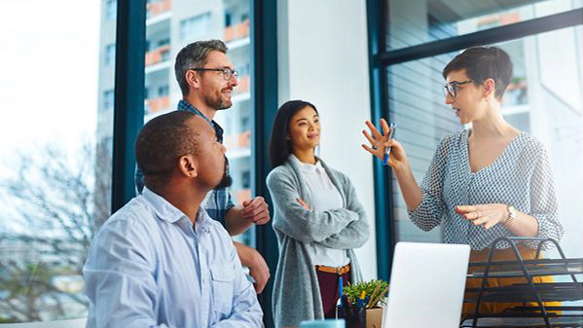 A group of professionals engaged in a discussion in a bright, modern office space. Large windows provide natural light and a view of urban buildings outside. The scene conveys teamwork and productivity, with a laptop visible on the desk. The atmosphere is professional and dynamic.