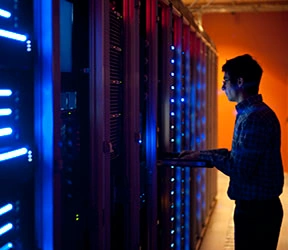The interior of a modern server room in a data center.  An IT engineer is busy configuring the servers. The room is dark, but the servers themselves are lit.  The servers at the left side are lit in blue, while the ones at the other end are lit by an orange light source coming from the right side.