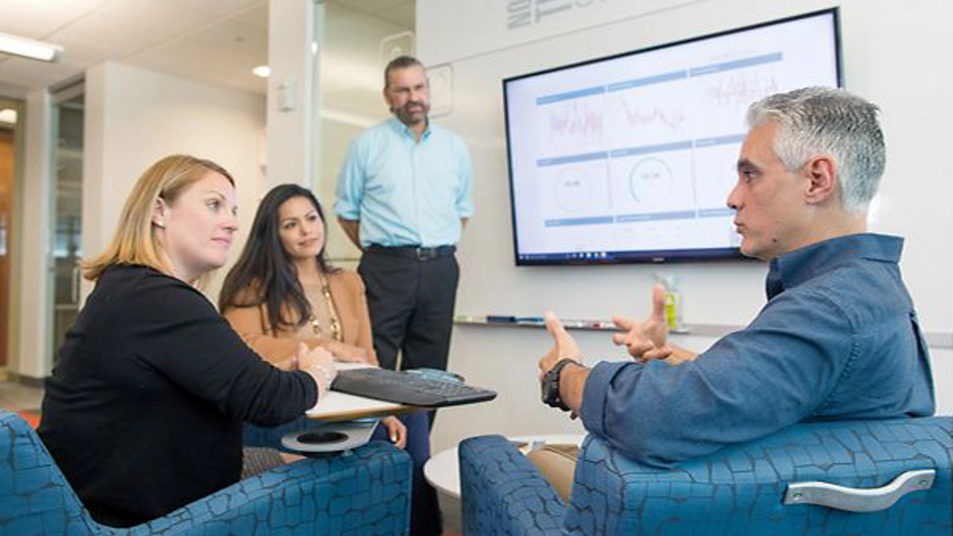 A group of professionals engaged in a meeting in a modern office setting. The focus is on a large presentation screen displaying graphs and charts, suggesting data analysis or business strategy discussion. The environment includes comfortable seating and a collaborative atmosphere.