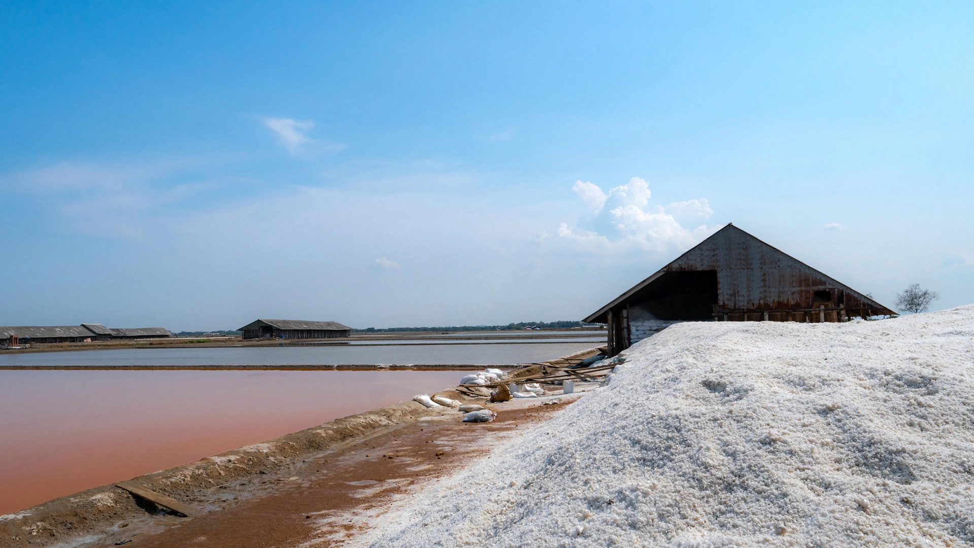 Landscape of brine salt farm with blue sky on sunny day. Salt warehouse. Pile of organic sea salt. Raw material of salt industrial. Sodium chloride mineral. Evaporation of sea water. Iodine.