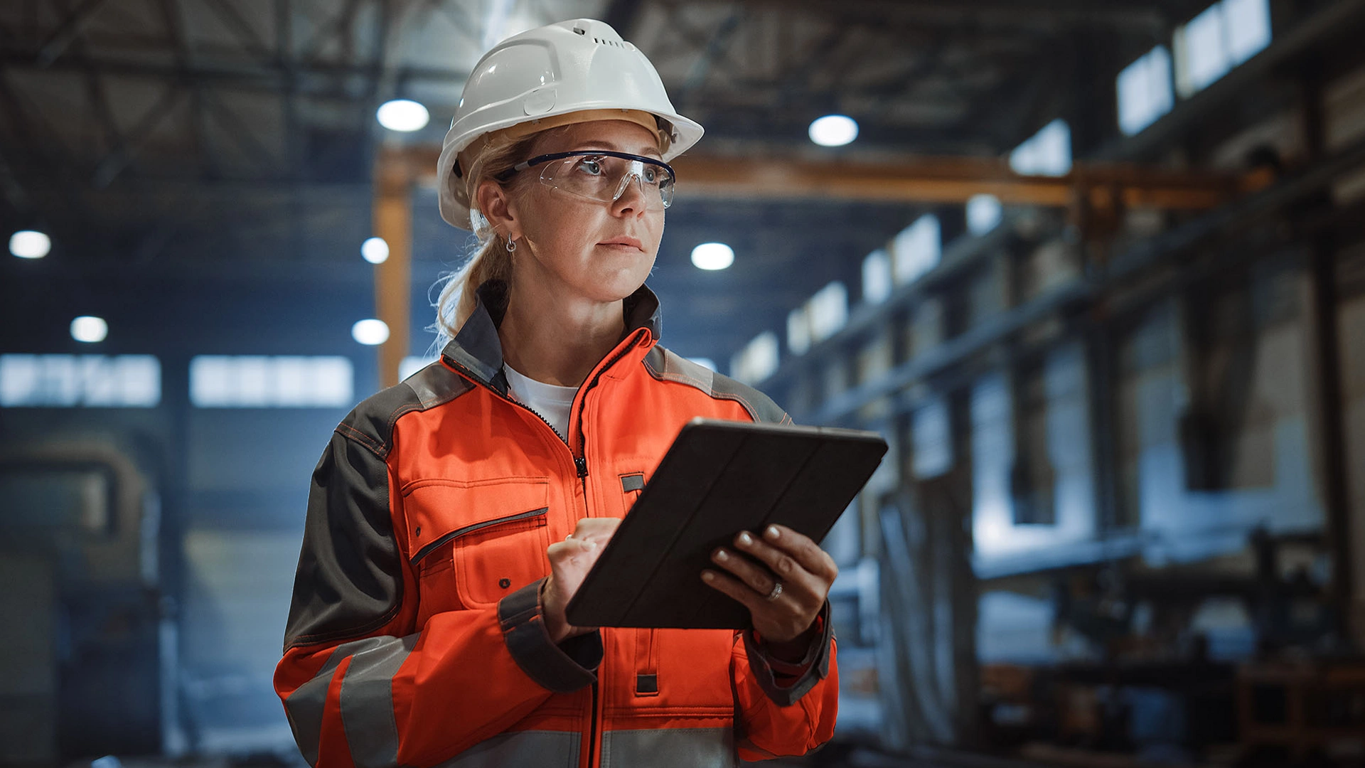 Professional Heavy Industry Engineer/Worker Wearing Safety Uniform and Hard Hat Uses Tablet Computer. Serious Successful Female Industrial Specialist Walking in a Metal Manufacture Warehouse.